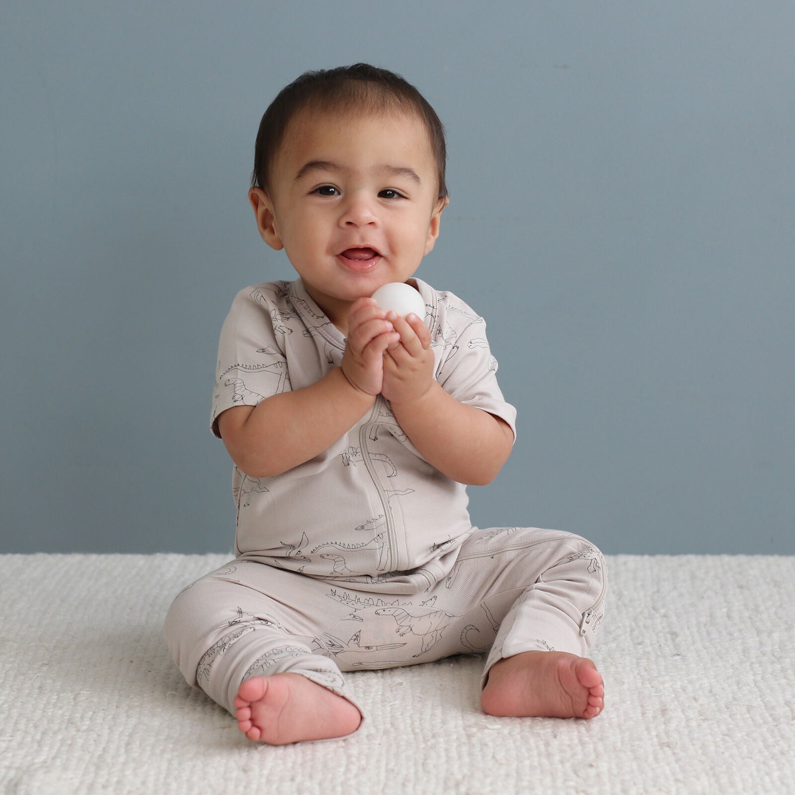 Baby sitting on a white blanket holding a white ball against a gray background