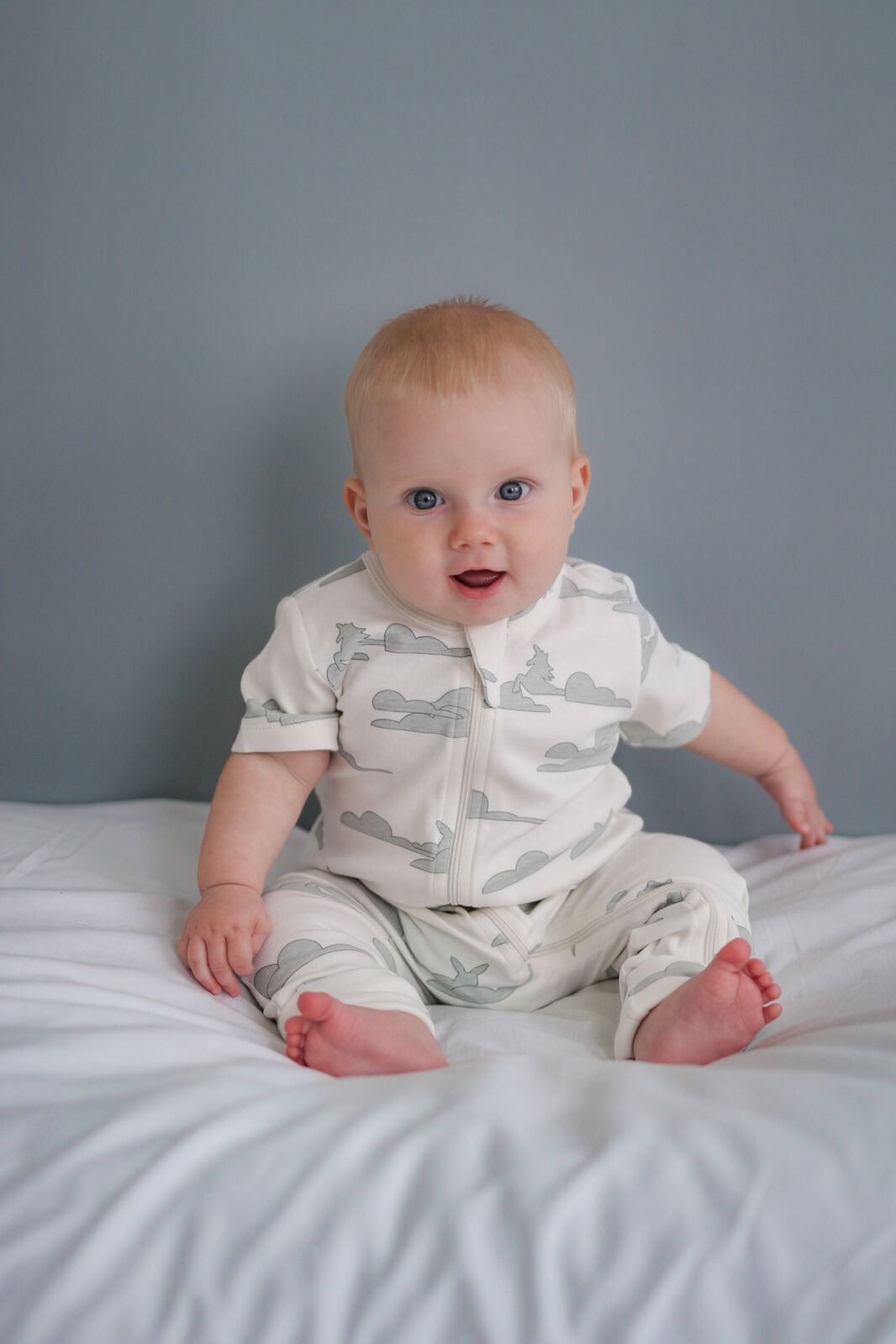Baby sitting on a bed wearing a white onesie with gray animal prints against a gray wall.
