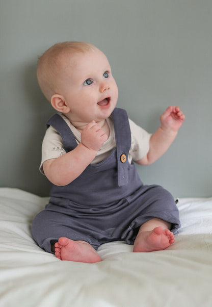 Baby wearing a purple romper sitting on a bed with a gray wall background