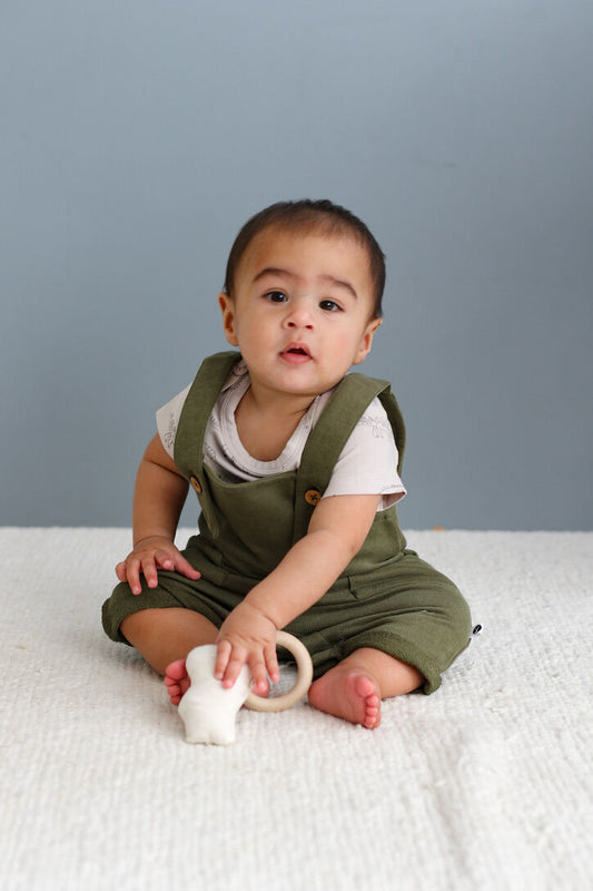 Baby in green overalls sitting on a white blanket with a gray background