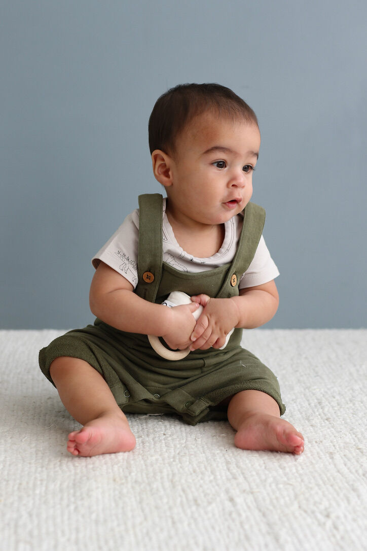 Baby wearing green overalls sitting on a white surface with a gray background
