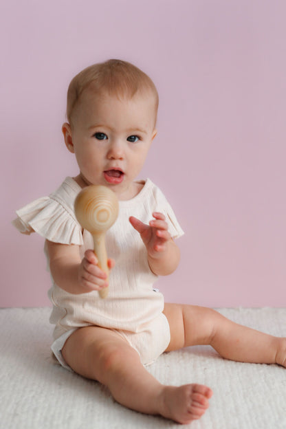 Baby in a white outfit holding a wooden rattle against a pink background