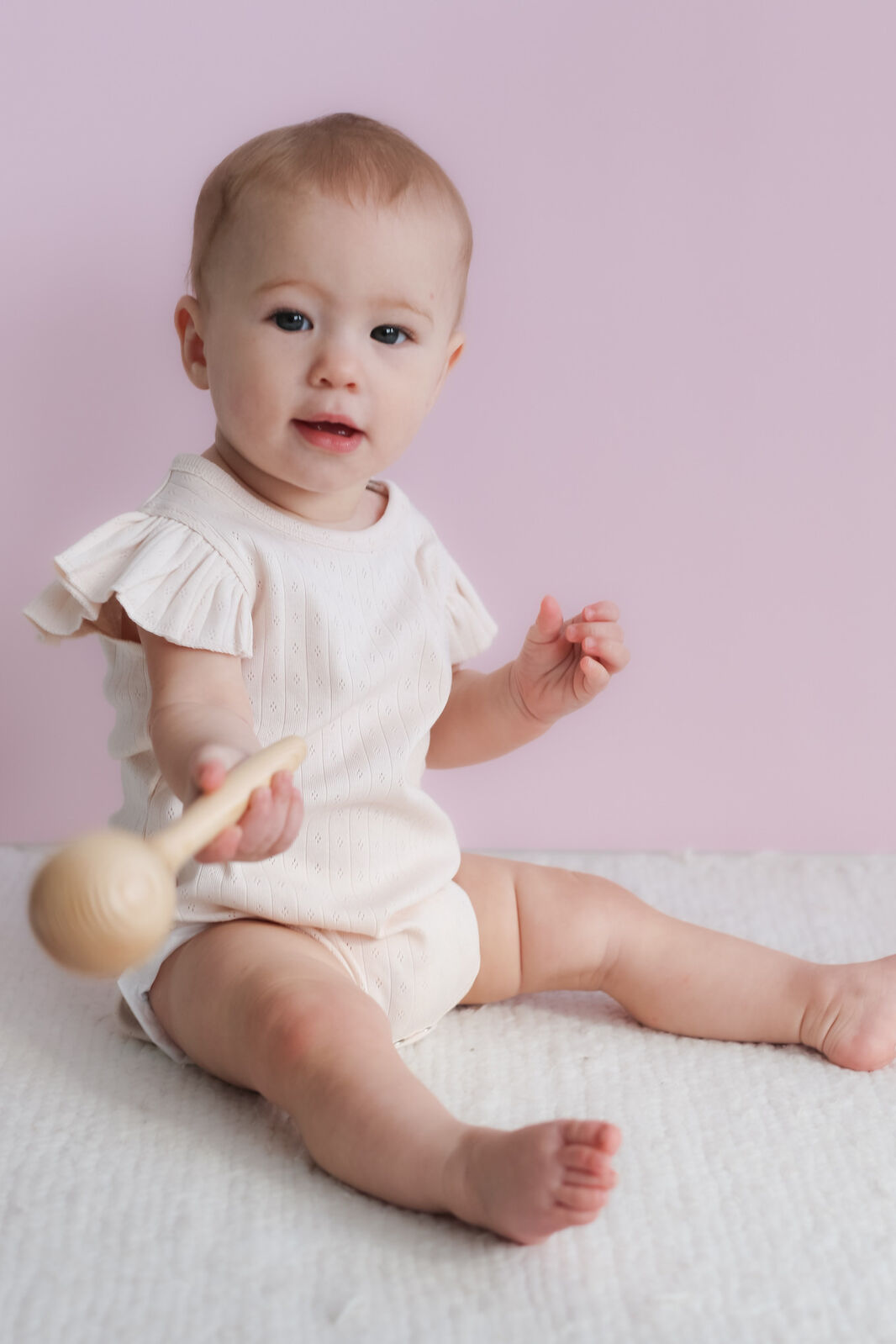 Baby in a white romper holding a wooden rattle on a light pink background