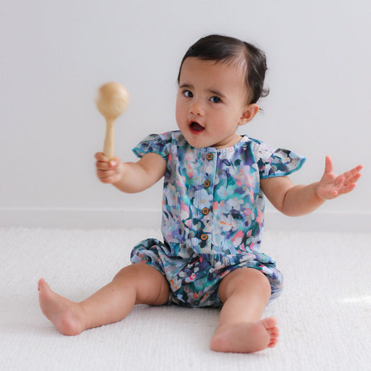 Baby in a floral romper holding a wooden rattle on a white background