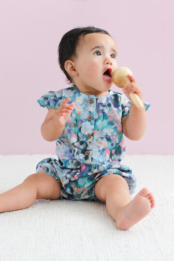 Baby in a floral outfit holding a wooden rattle against a light pink background