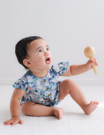 Baby in a floral onesie holding a wooden rattle on a white background