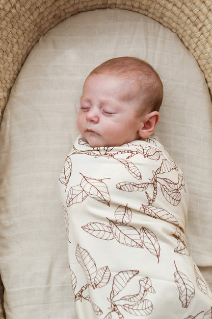 Baby laying in Moses Basket, wrapped in Leaves Stretchy Swaddle, designed by Burrow and Be