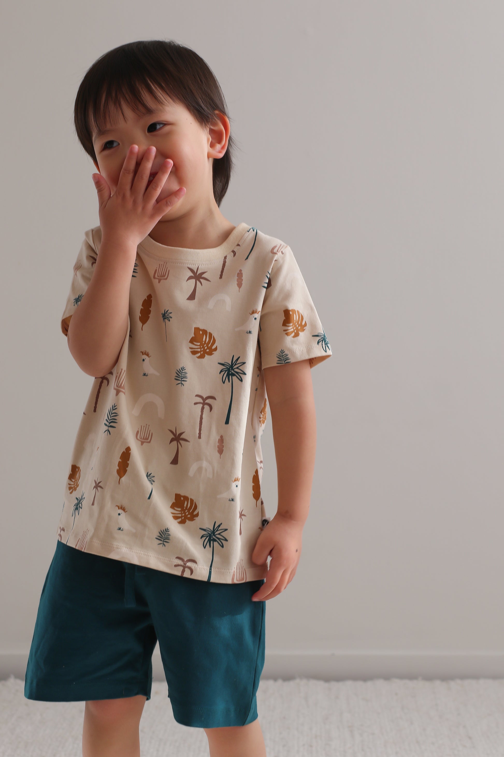 Boy laughing at camera while standing against a white wall and wearing Deep Teal Leo Shorts and Jungle Stamp Classic T-Shirt, designed by Burrow and Be