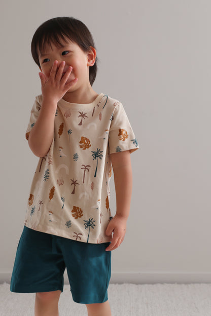Boy laughing at camera while standing against a white wall and wearing Deep Teal Leo Shorts and Jungle Stamp Classic T-Shirt, designed by Burrow and Be
