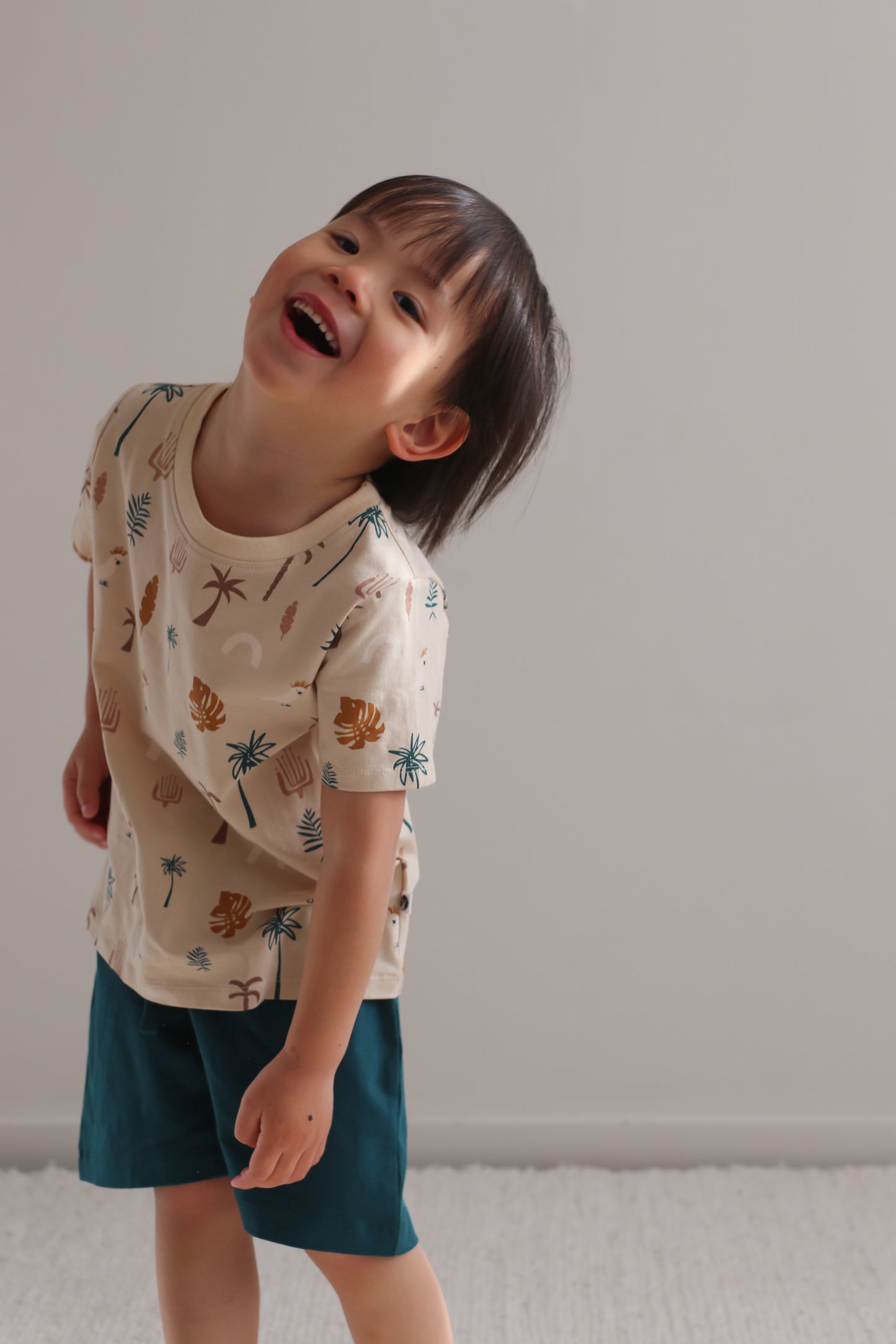 Boy laughing at camera while standing against a white wall and wearing Leo Shorts in Deep Teal and Jungle Stamp Classic T-Shirt, all designed by Burrow and Be