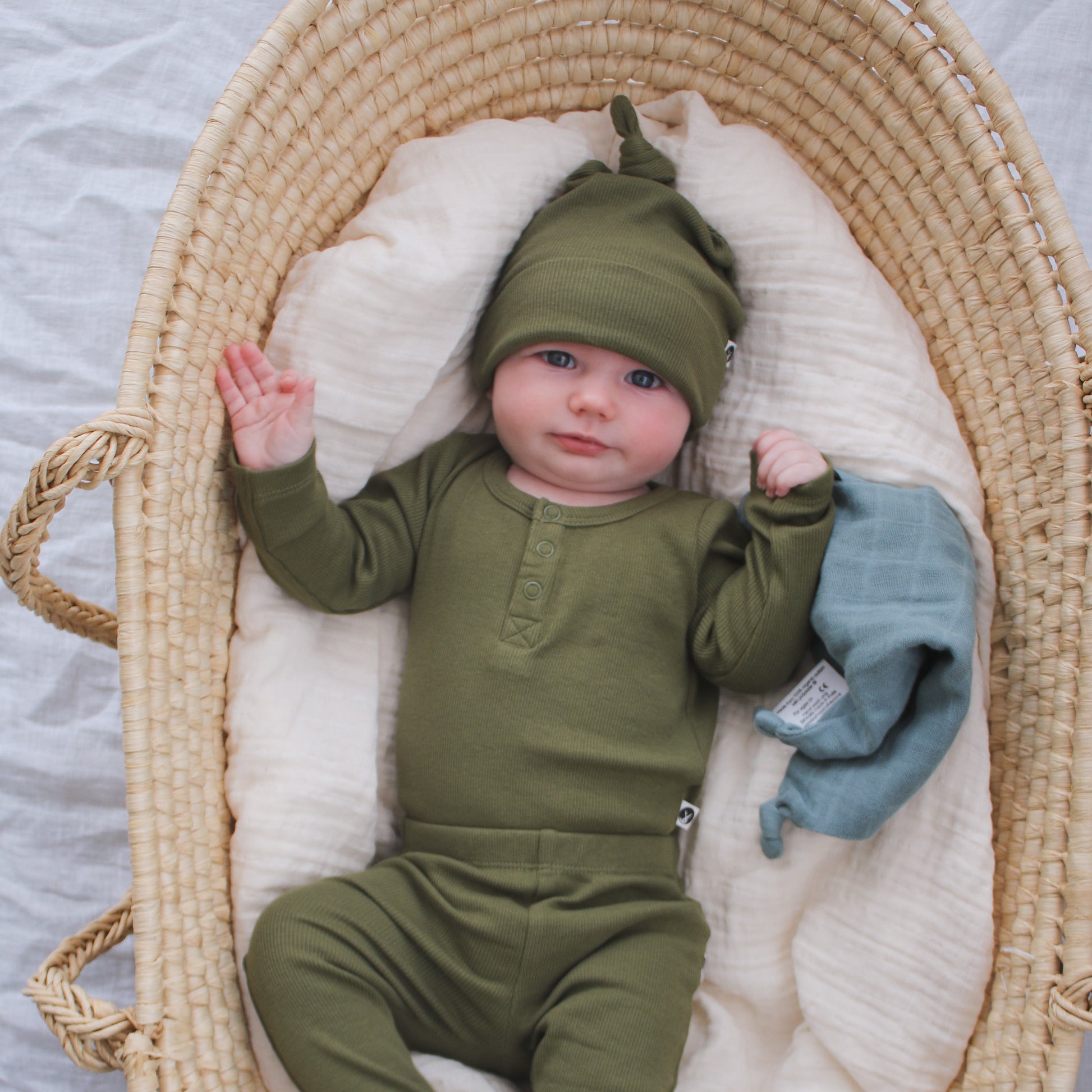 Baby laying in Moses Basket with Dusty Rose Muslin while wearing Long Sleeve Henley Rib Bodysuit, Leggings and Beanie all in Olive and designed by Burrow and Be