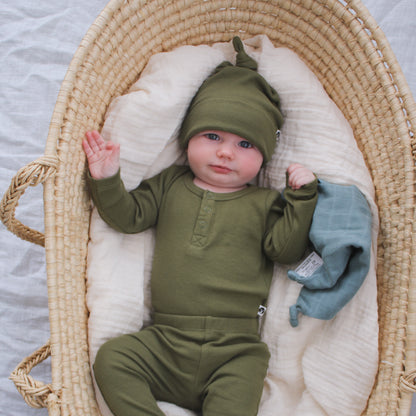 Baby laying in Moses Basket with Dusty Rose Muslin while wearing Long Sleeve Henley Rib Bodysuit, Leggings and Beanie all in Olive and designed by Burrow and Be