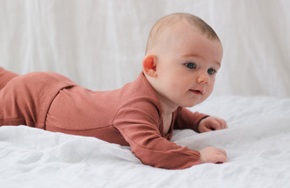 Baby laying on white muslin sheets while wearing a Long Sleeve Henley Rib Bodysuit and Leggings in Clay, designed by Burrow and Be