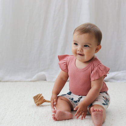 Girl sitting on the floor in front of white curtains and wearing Pointelle Flutter Bodysuit in Dusk and Pink Clementine Bike Shorts, both designed by Burrow and Be