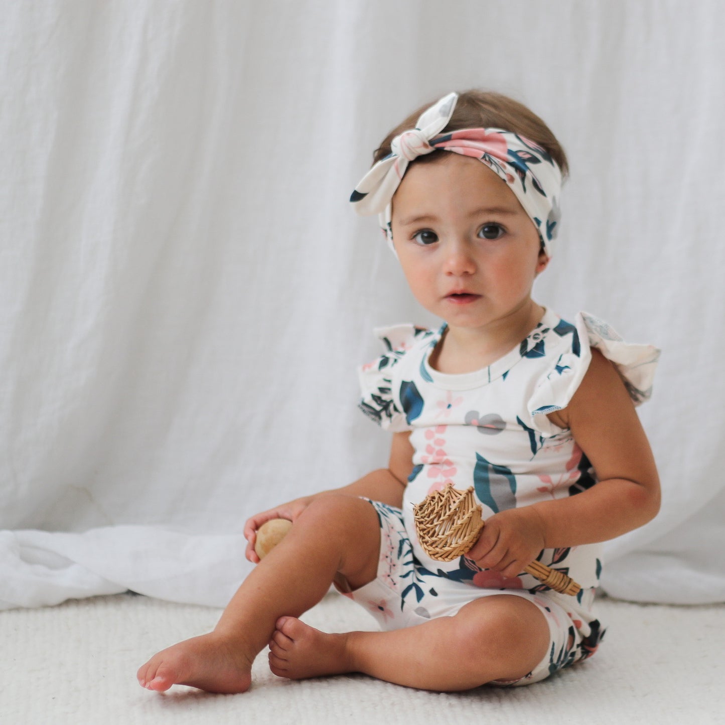 Girl sitting on the carpet in front of white curtains and wearing Pink Clementine Headband, Flutter Bodysuit and Bike Shorts, all designed by Burrow and Be