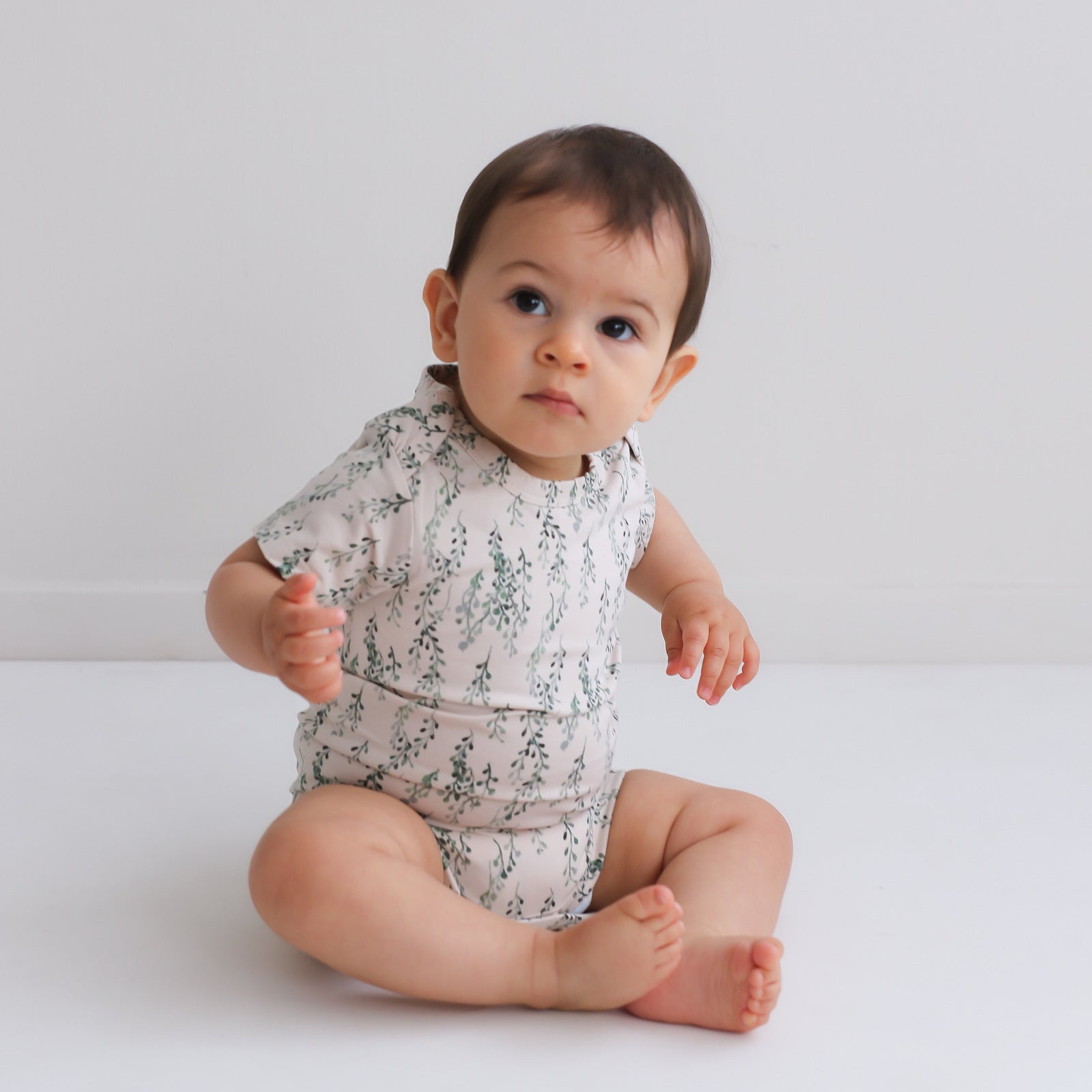 Baby sitting on white floor in front of a white wall while wearing String of Pearls Short Sleeve Bodysuit, designed by Burrow and Be