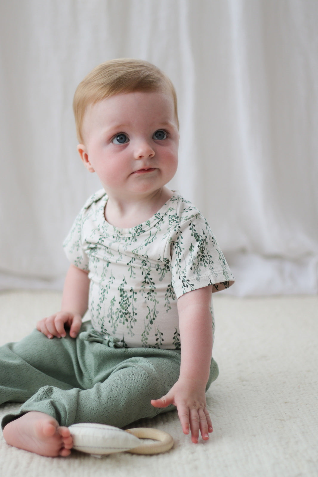 Baby siting on carpet in front of a white curtain while wearing Pointelle Baby Pants in Sage and String of Pearls Short Sleeve Bodysuit, designed by Burrow and Be