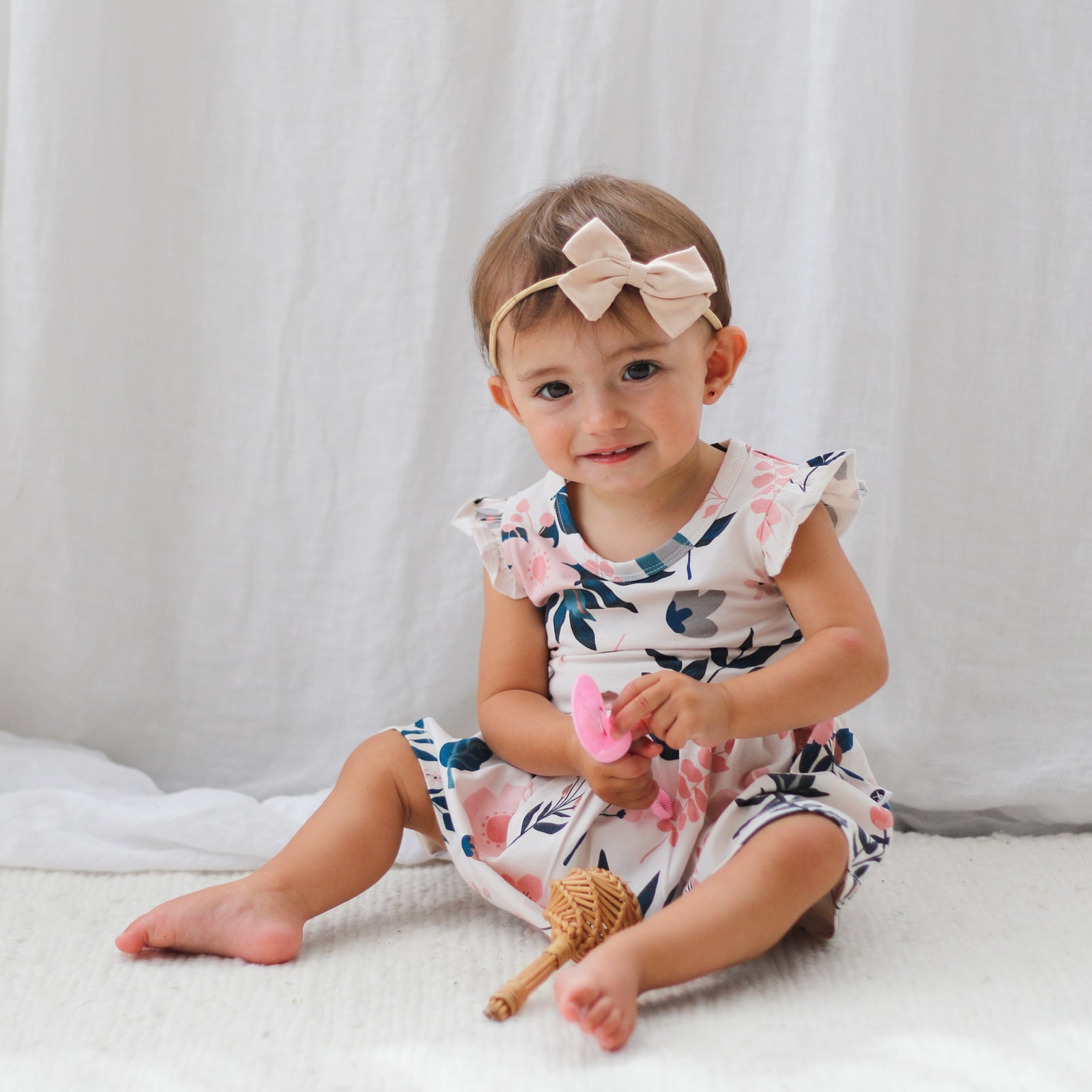 Baby sitting on carpet in front of white curtains and wearing Bow Tie Hair Tie and Pink Clementine Flutter Baby Dress, all designed by Burrow and Be