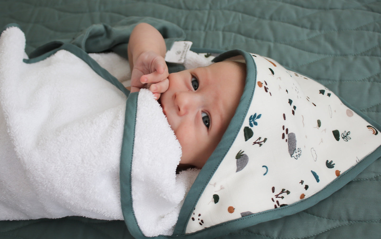Baby posing at camera while laying on a Sage quilted mattress and wrapped in Garden Treasures Baby Hooded Towel, all designed by Burrow and Be