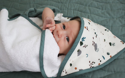 Baby posing at camera while laying on a Sage quilted mattress and wrapped in Garden Treasures Baby Hooded Towel, all designed by Burrow and Be
