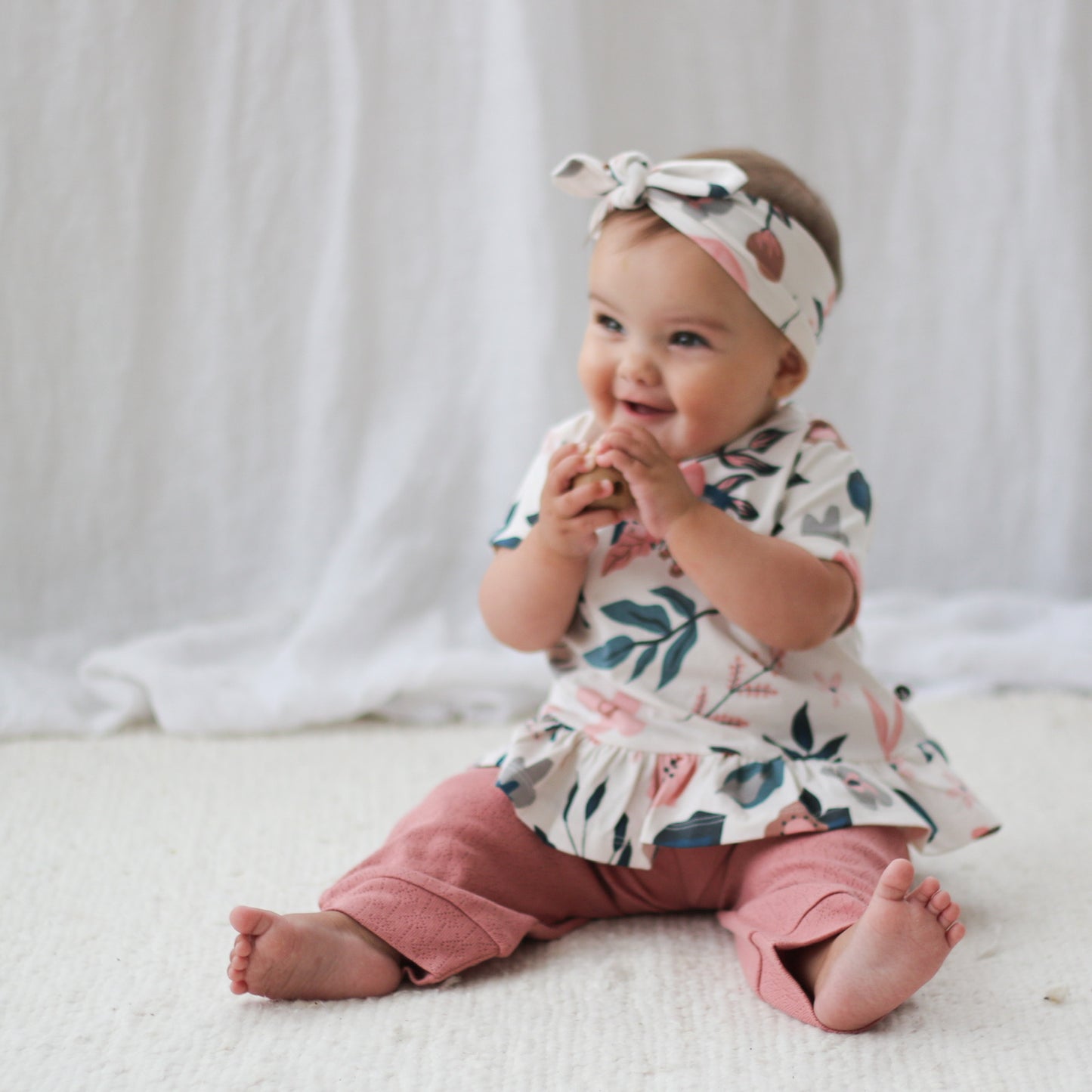 Baby sitting on a carpet in front of a white curtain while wearing Pink Clementine Headband, Hana Top and Pointelle Long Pants in Dusk, all designed by Burrow and Be