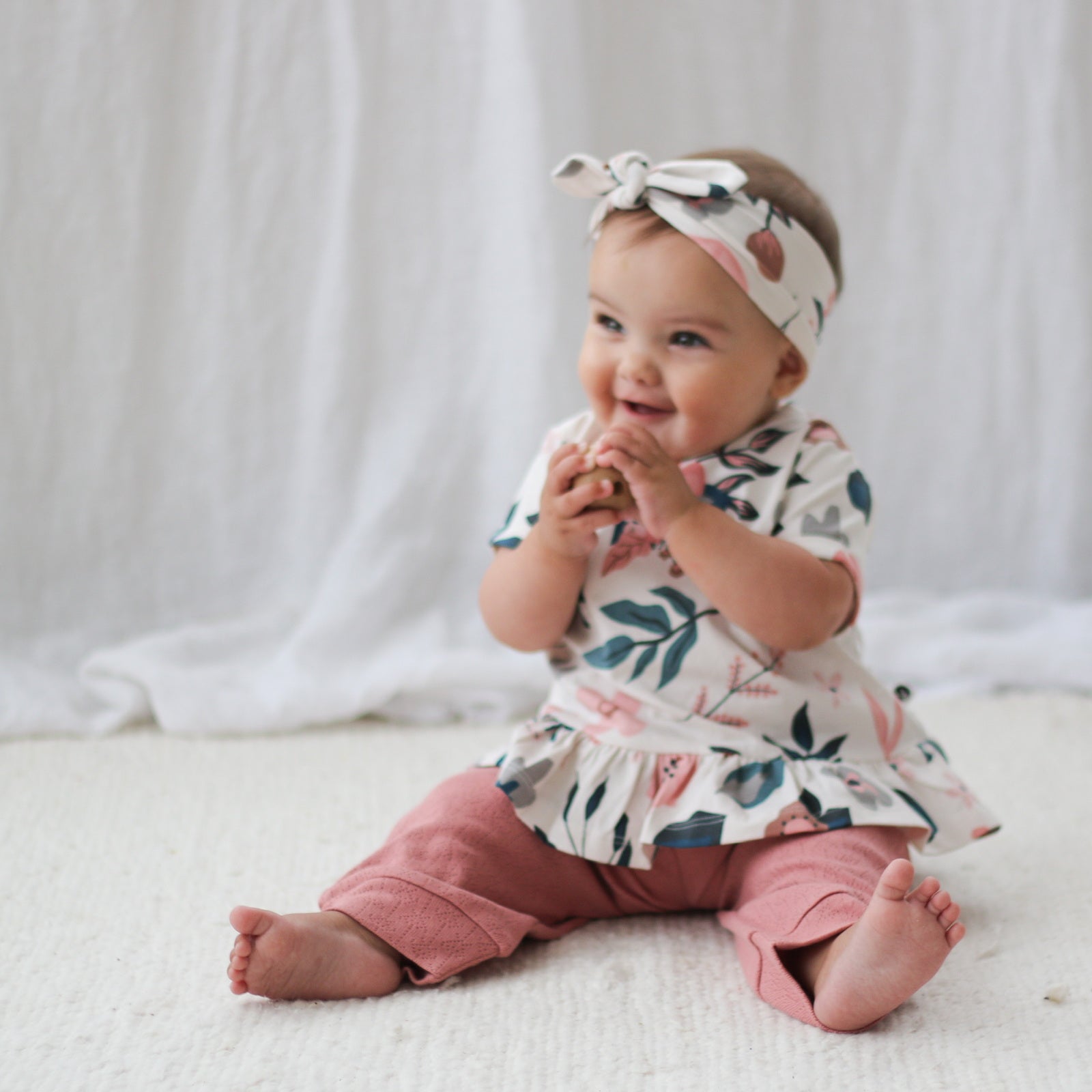 Baby sitting on a carpet in front of a white curtain while wearing Pink Clementine Headband, Hana Top and Pointelle Long Pants in Dusk, all designed by Burrow and Be