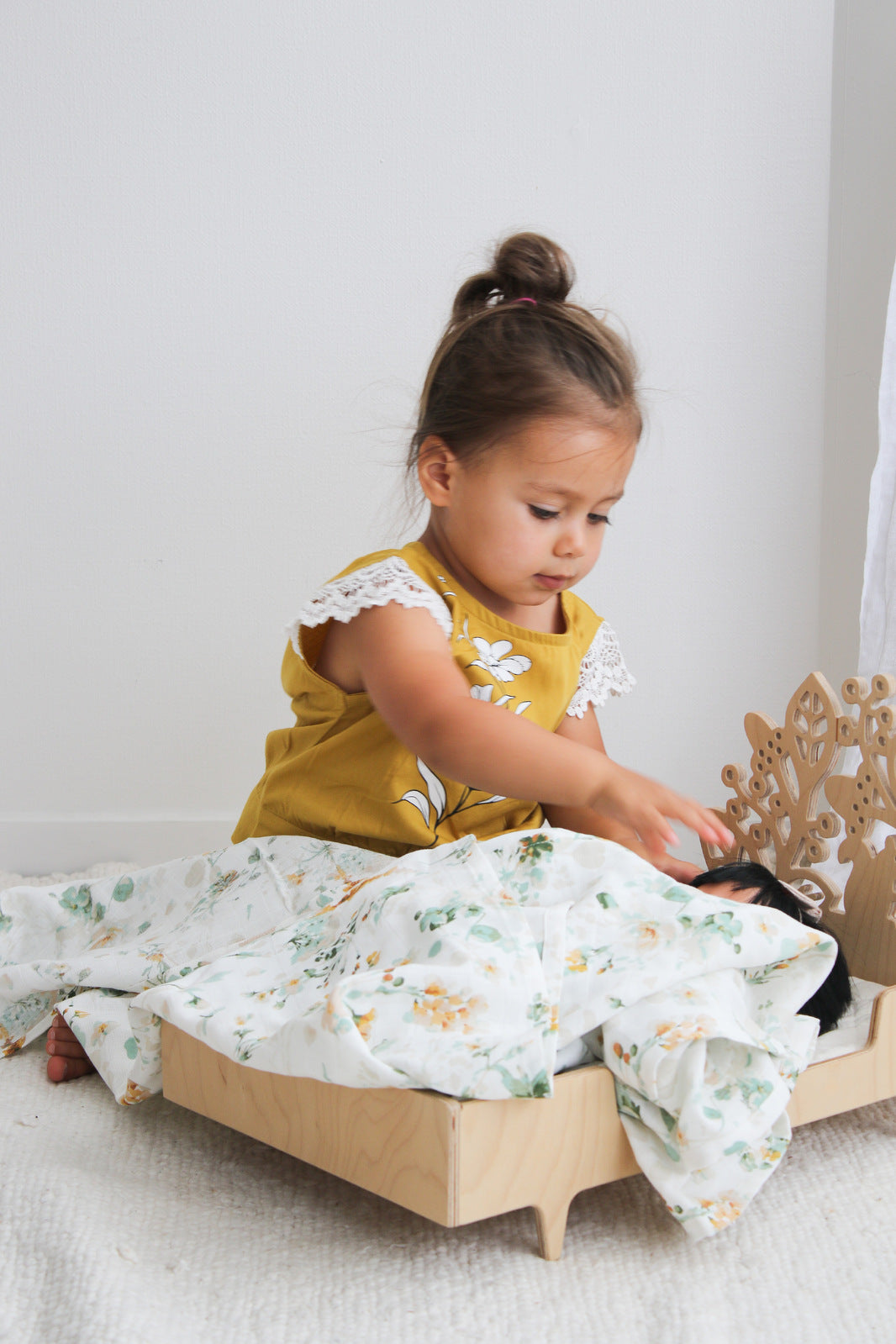 Girl playing with dolls in front of a white wall, wrapping the doll in Spring Melody Muslin Wrap, designed by Burrow and Be