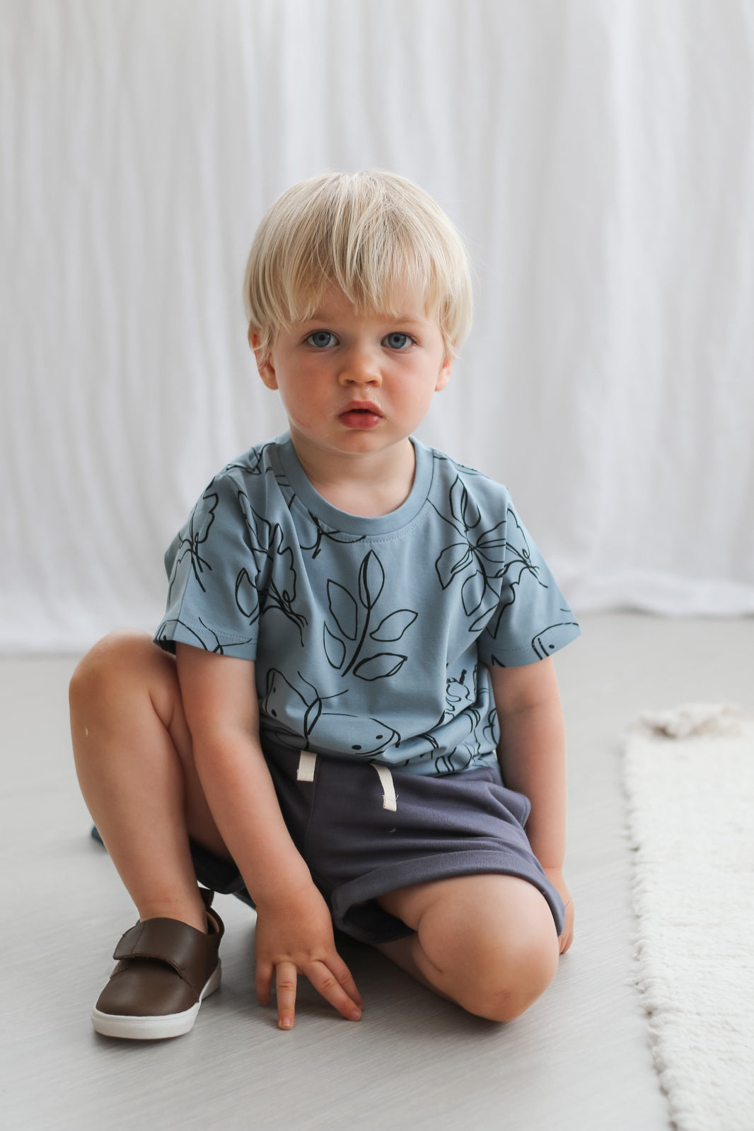 Boy posing in front of the camera wearing Shoes by Wanders, Giant Bugs T-shirt and Terry Knit Baby Shorts in Ink Blue against white curtains, all designed by Burrow and Be