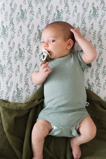 Baby laying on Strings of Pearl Sheets and Essential Muslin Wrap in Pine, using Pine Essential Pacifier Clip while posing in Pointelle Short Sleeve Bodysuit in Riverstone, all designed by Burrow and Be