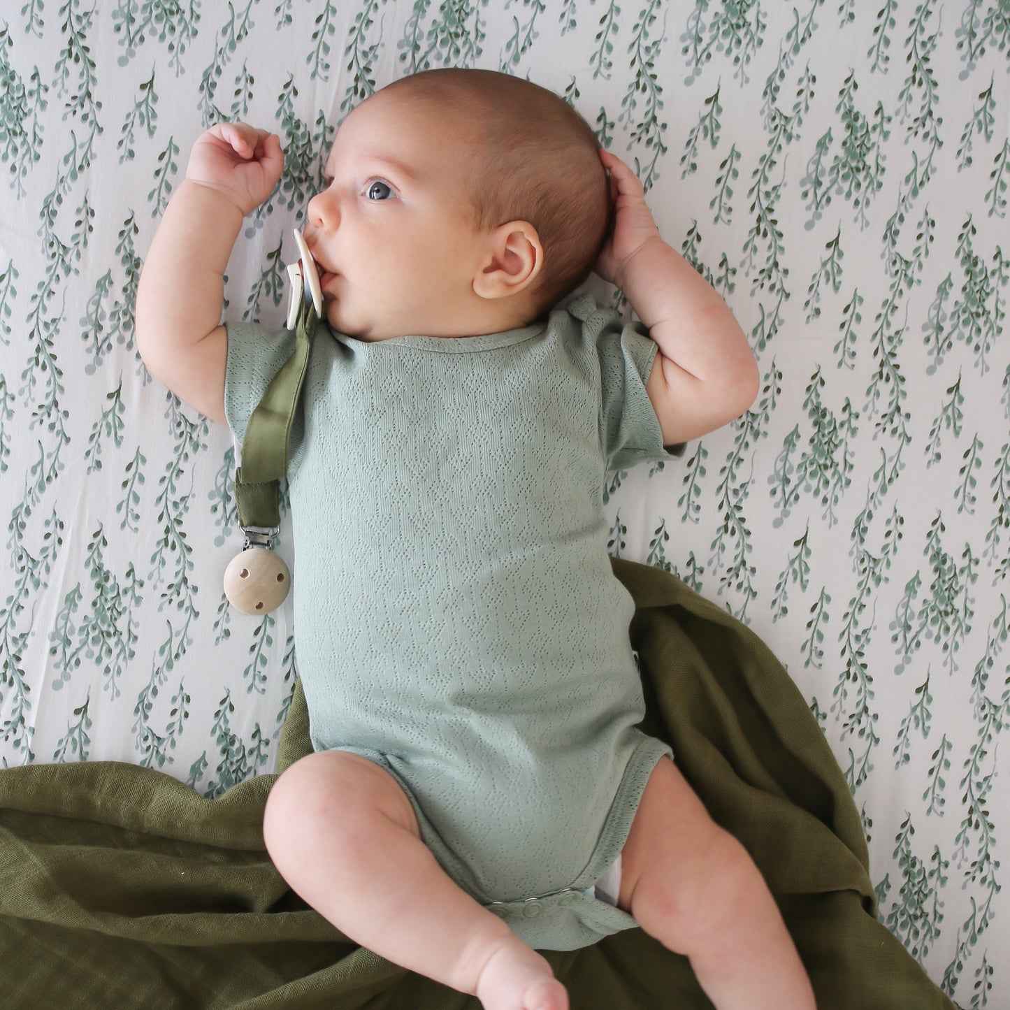 Baby laying on Strings of Pearl Sheets and Essential Muslin Wrap in Pine, using Pine Essential Pacifier Clip while wearing Pointelle Short Sleeve Bodysuit in Riverstone, all designed by Burrow and Be
