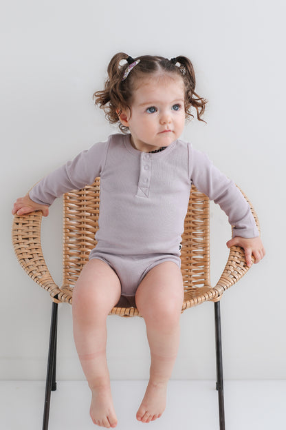 Baby sitting on a chair against a white background while wearing a Long Sleeve Henley Rib Bodysuit in Lavender, designed by Burrow and Be