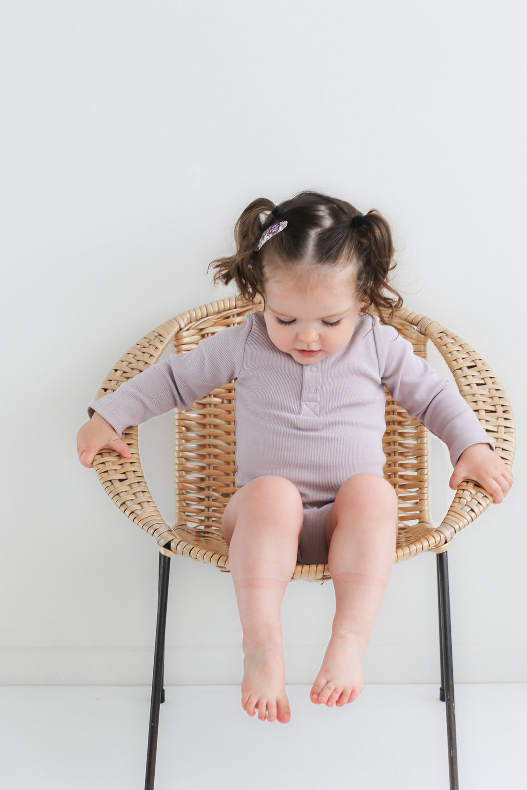 Baby sitting on a chair against a white background looking at the ground while wearing a Long Sleeve Henley Rib Bodysuit in Lavender, designed by Burrow and Be