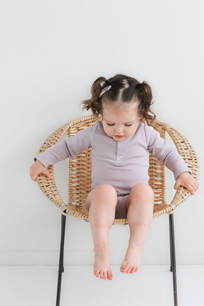 Baby sitting on a chair against a white background looking at the ground while wearing a Long Sleeve Henley Rib Bodysuit in Lavender, designed by Burrow and Be