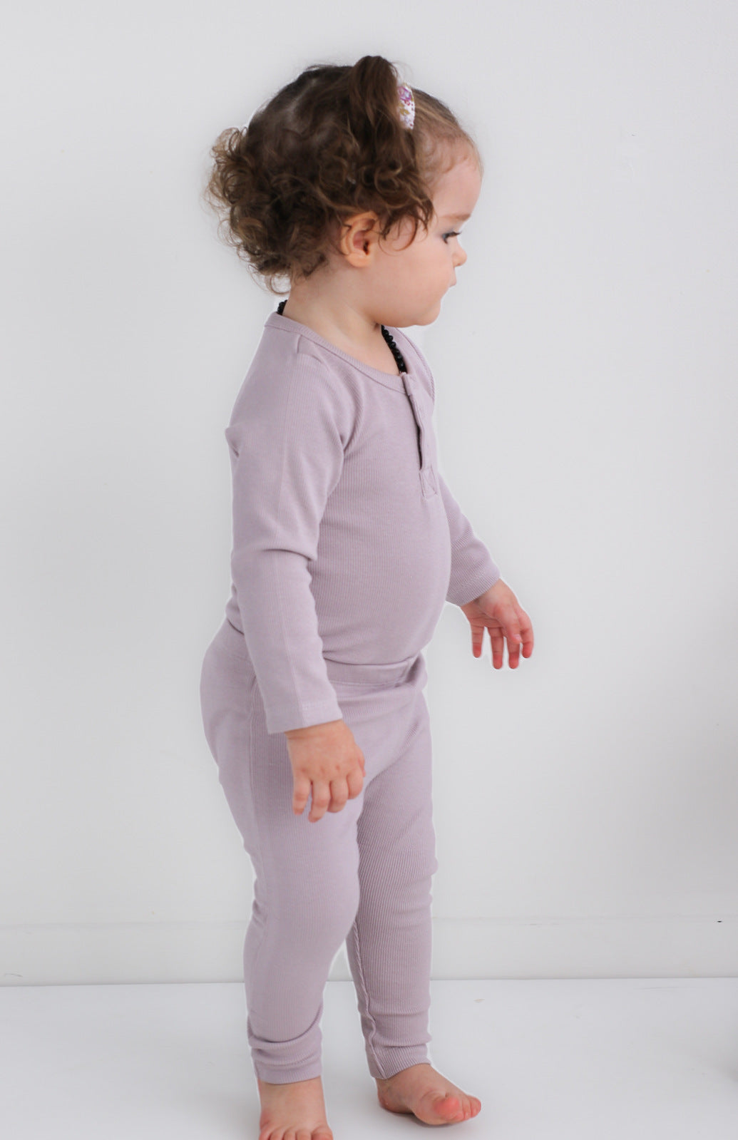 Baby standing against a white background while wearing a Long Sleeve Henley Rib Bodysuit and Leggings in Lavender, all designed by Burrow and Be