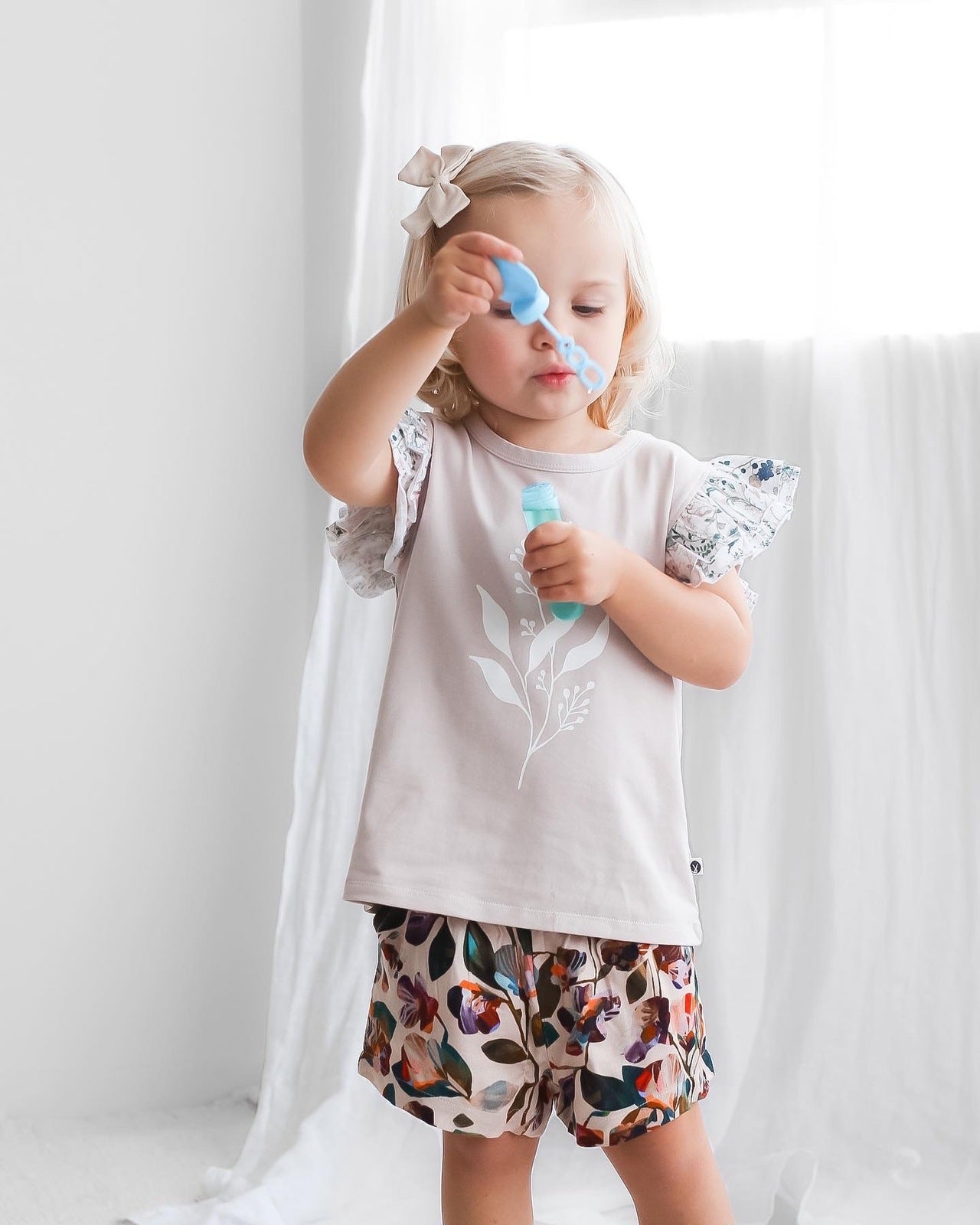 Girl blowing bubbles while wearing Wildflower/Lavender Flutter T-shirt and Garden Party Lyla Shorts by Burrow and Be