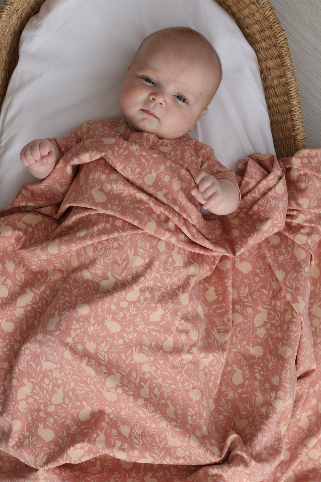 Baby lying in a Moses Basket, wearing a Mead-Doe Zip Suit and wrapped in Stretchy Swaddle, all designed by Burrow and Be