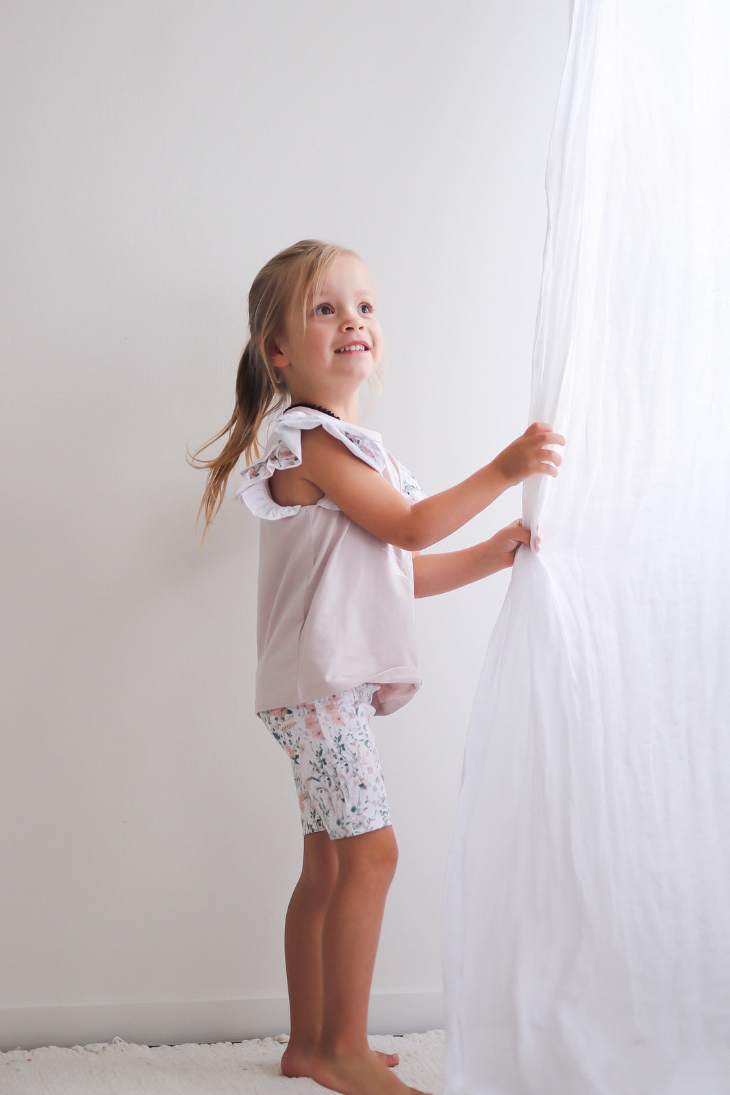 Girl standing in front of white background and playing with white curtains, wearing Wildflower/Lavender Flutter T-Shirt and Wildflower Bike Shorts, both designed by Burrow and Be