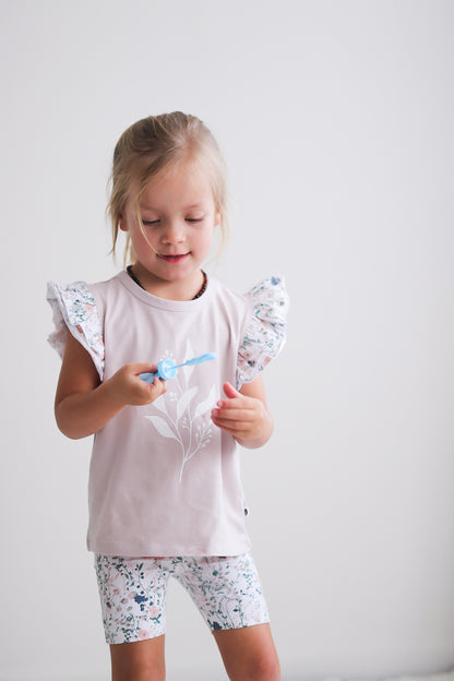 Girl standing in front of white background and playing with bubbles, wearing Wildflower/Lavender Flutter T-Shirt and Wildflower Bike Shorts, both designed by Burrow and Be