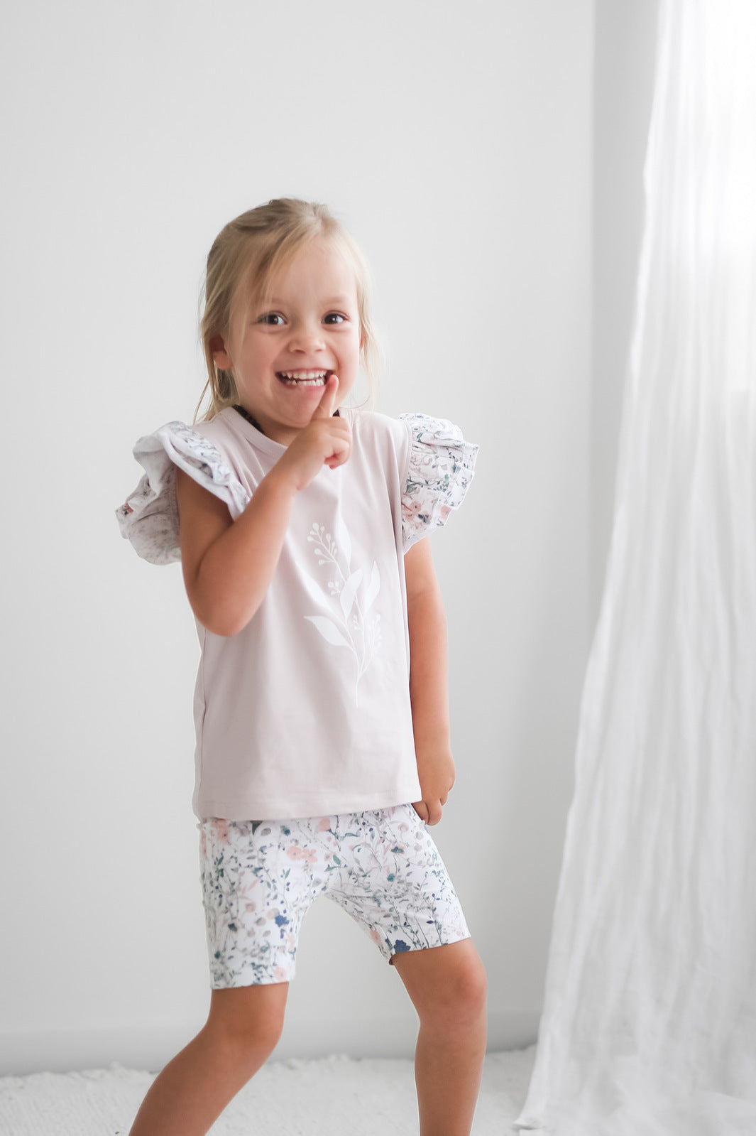 Girl standing in front of white background, posing at the camera wearing Wildflower/Lavender Flutter T-Shirt and Wildflower Bike Shorts, both designed by Burrow and Be