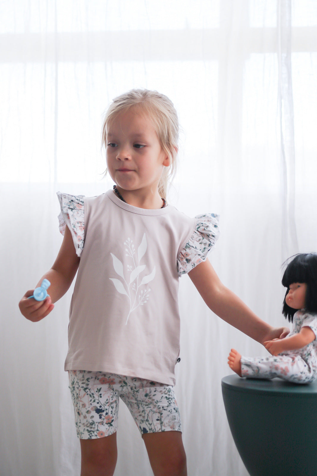 Girl standing in front of white curtains, posing with doll wearing Wildflower Doll Clothes and wearing Wildflower/Lavender Flutter T-Shirt and Wildflower Bike Shorts, both designed by Burrow and Be