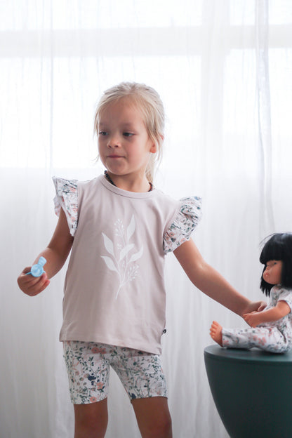 Girl standing in front of white curtains, posing with doll wearing Wildflower Doll Clothes and wearing Wildflower/Lavender Flutter T-Shirt and Wildflower Bike Shorts, both designed by Burrow and Be