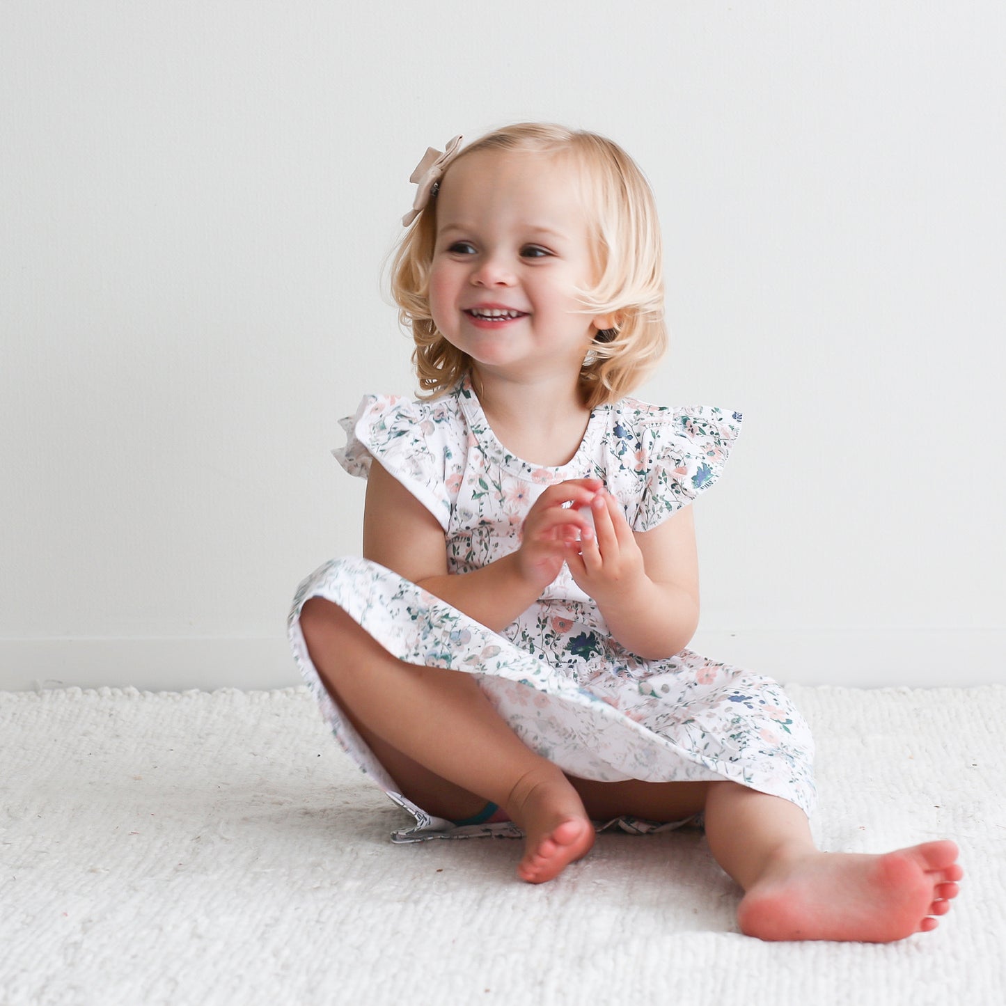 Girl sitting on carpet in front of a white background, wearing Wildflower Flutter Dress, designed by Burrow and Be