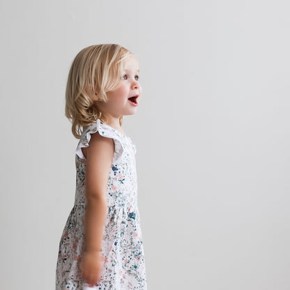 Girl standing in front of a white background facing left while wearing Wildflower Flutter Dress, designed by Burrow and Be