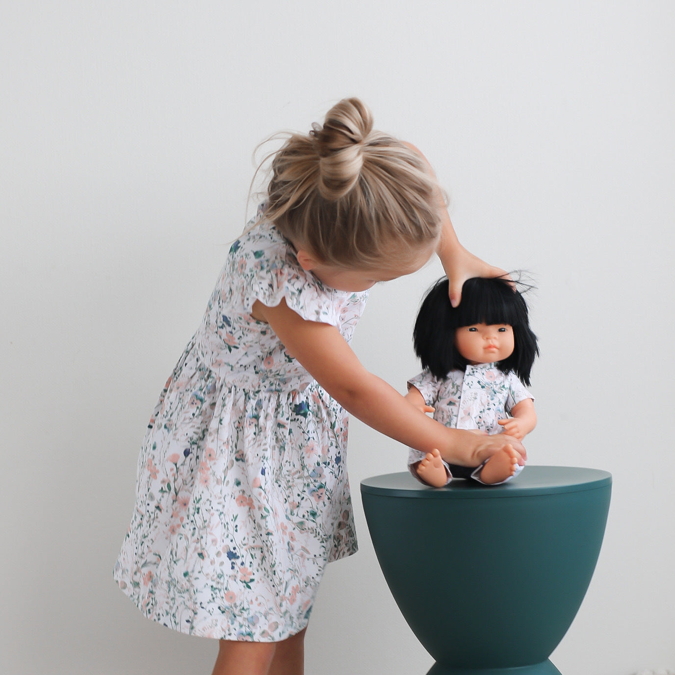Girl standing in front of a white background, playing with a doll seated on a stool wearing Wildflower Doll Clothes while wearing Wildflower Flutter Dress, designed by Burrow and Be