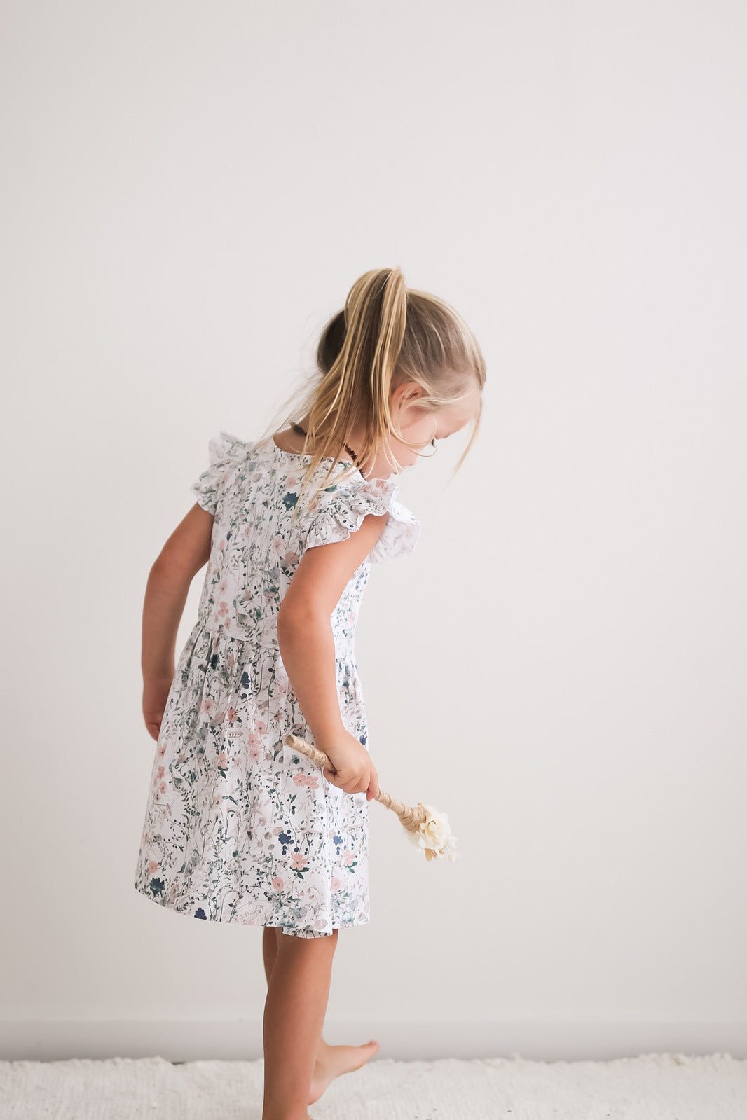 Girl standing facing a white wall while wearing Wildflower Flutter Dress, designed by Burrow and Be