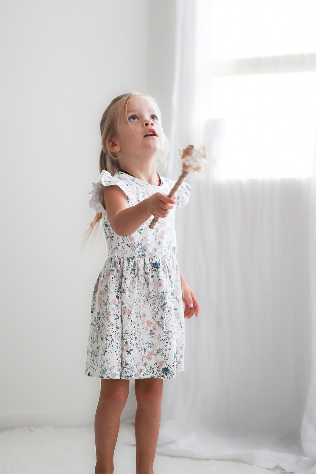 Girl standing in front of a white curtain while wearing Wildflower Flutter Dress, designed by Burrow and Be