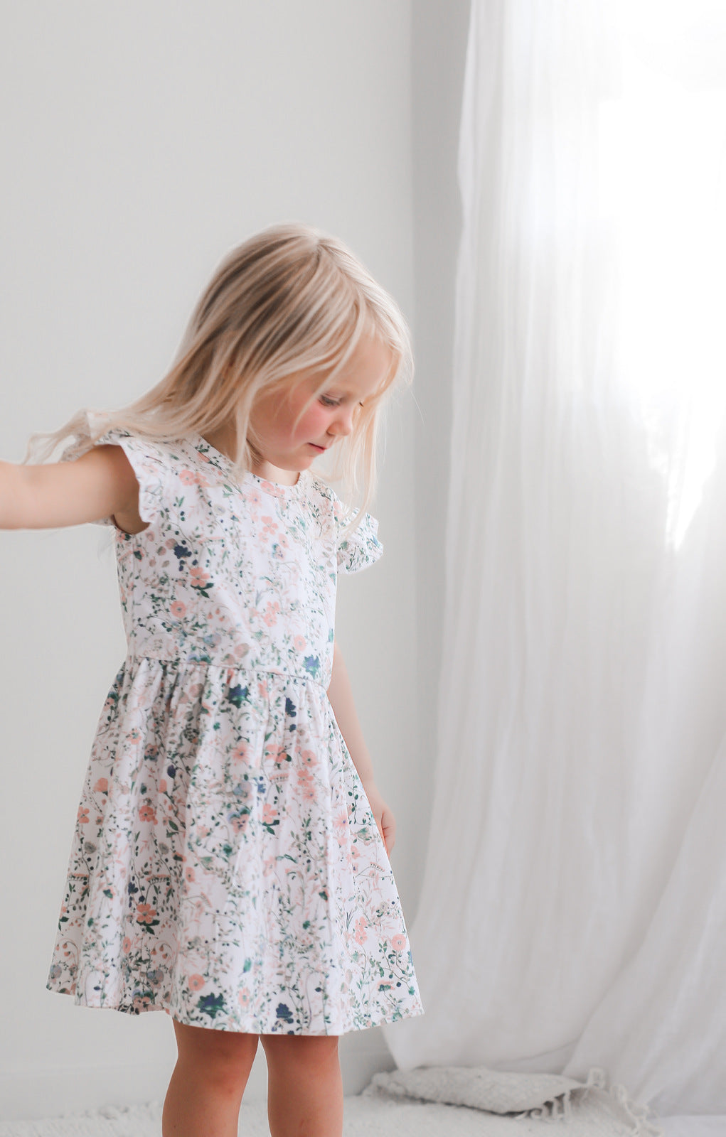 Girl standing in front of a white curtain while wearing Wildflower Flutter Dress, designed by Burrow and Be