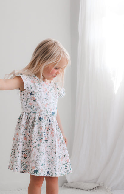 Girl standing in front of a white curtain while wearing Wildflower Flutter Dress, designed by Burrow and Be