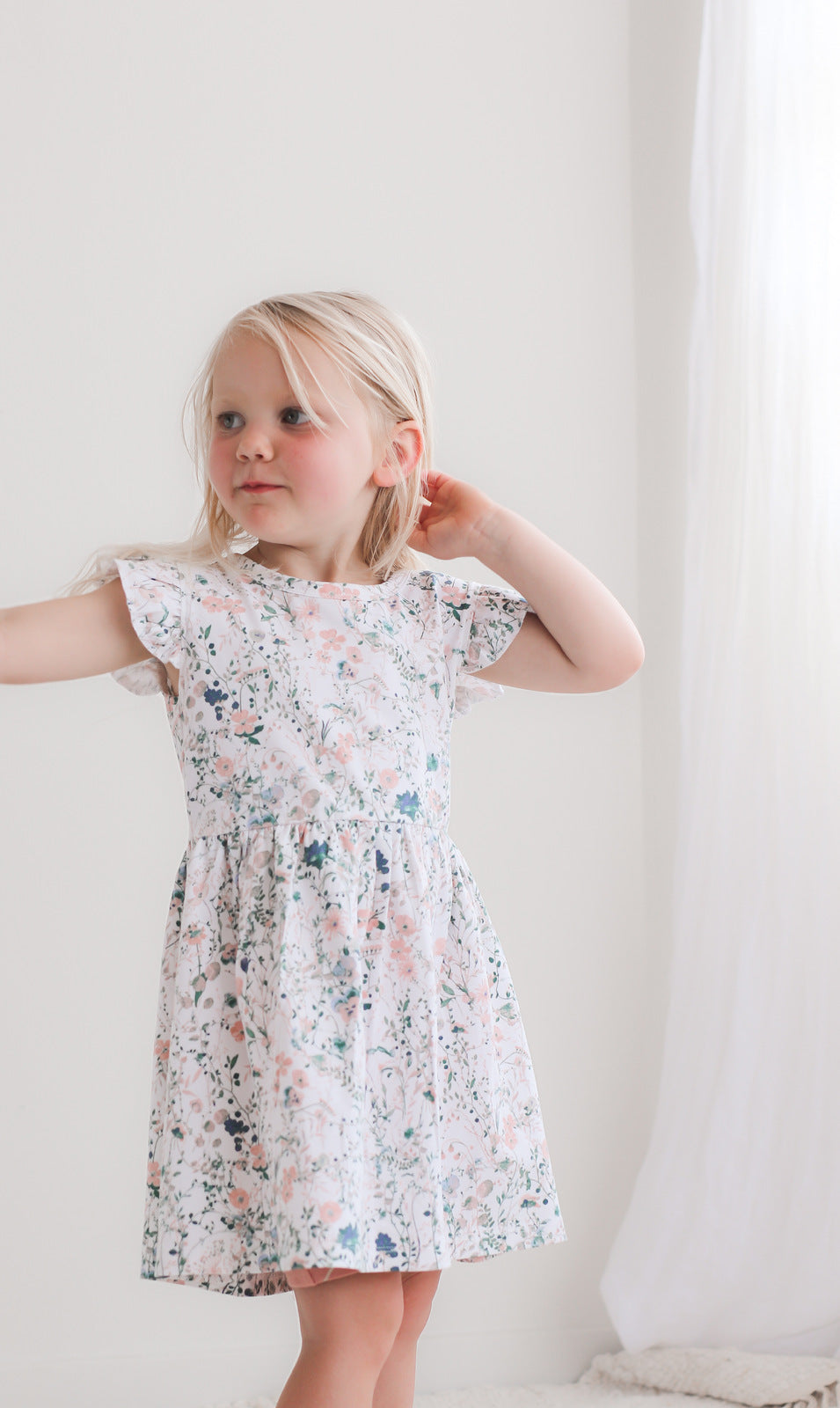 Girl standing in front of a white wall against curtain while wearing Wildflower Flutter Dress, designed by Burrow and Be