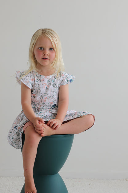 Girl sitting on a stool in front of a white wall while wearing Wildflower Flutter Dress, designed by Burrow and Be